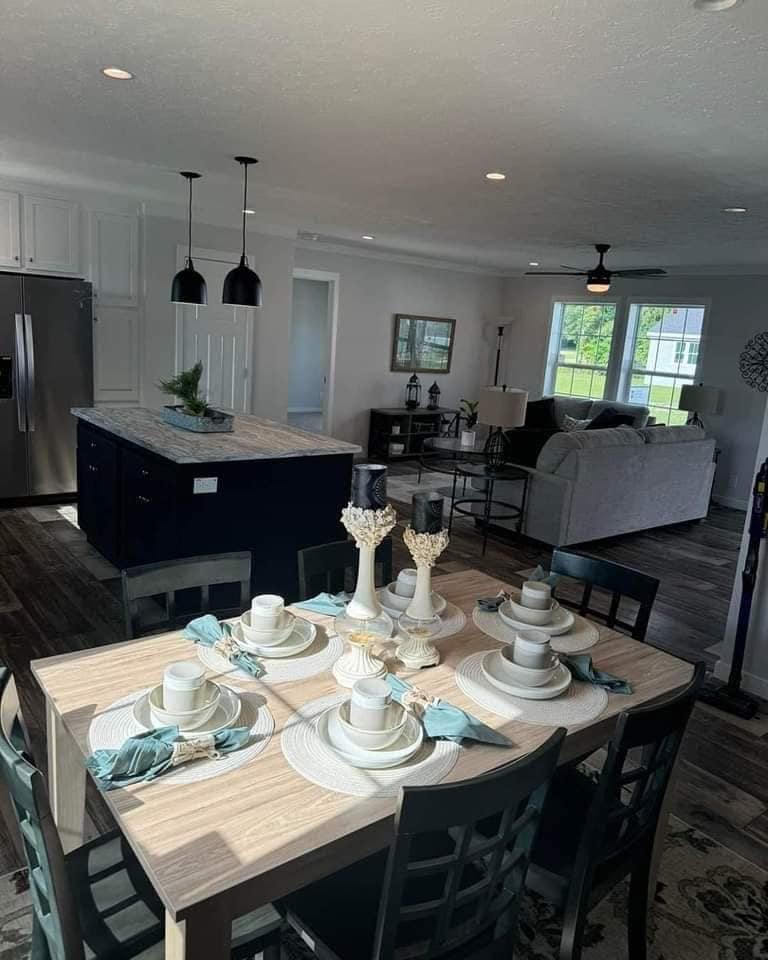 Modern dining and living area with a set dining table in the foreground. Kitchen island and stainless steel fridge on the left, couch and large windows in the background. Bright, open, and inviting space.
