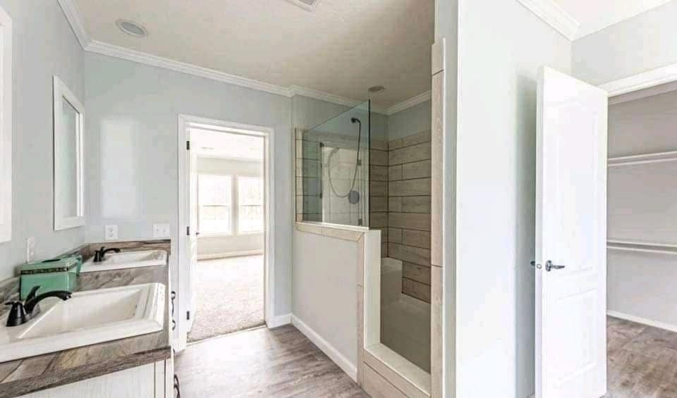 A modern bathroom with light gray walls features a double sink vanity with black faucets, a glass shower, and an open door leading to a bright bedroom.