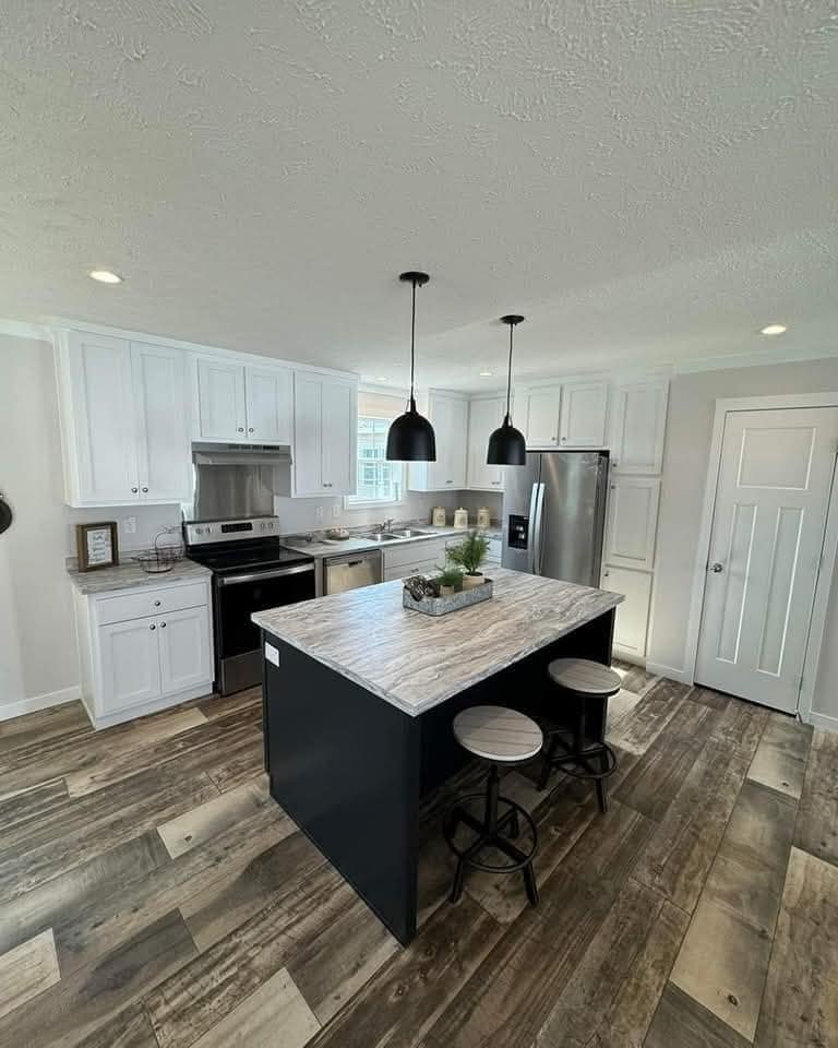 Modern kitchen with a marble island, two stools, dark pendant lights, and white cabinets. Stainless steel appliances and wood-look flooring. Bright and inviting atmosphere.