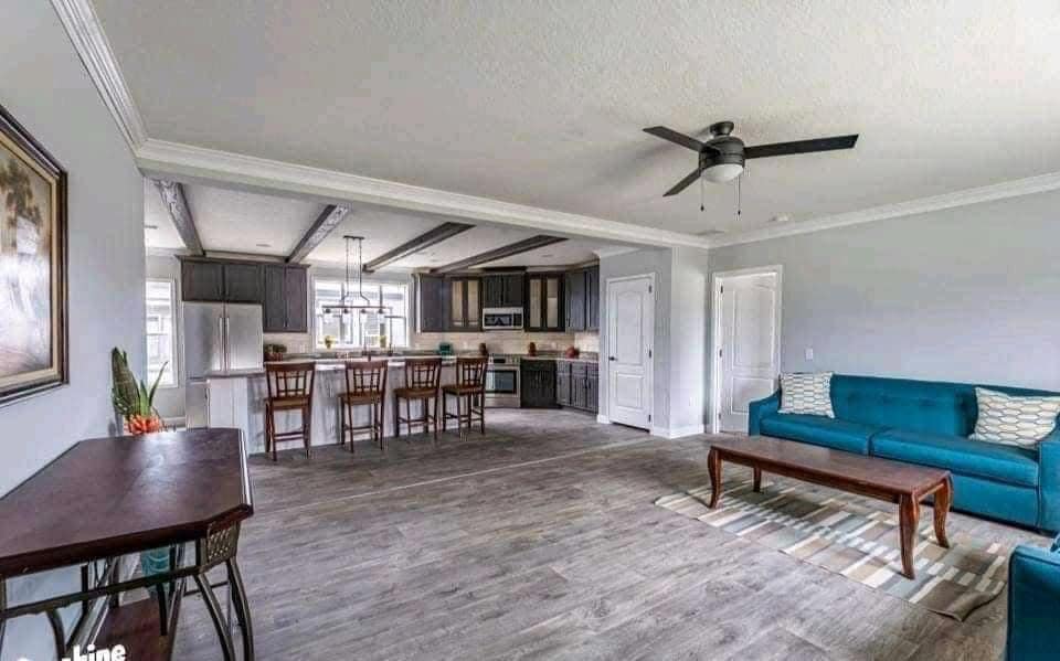 Spacious living area with gray wood flooring, teal sofa, and patterned rug. Open kitchen features barstools and stainless steel fridge. Bright and modern ambiance.