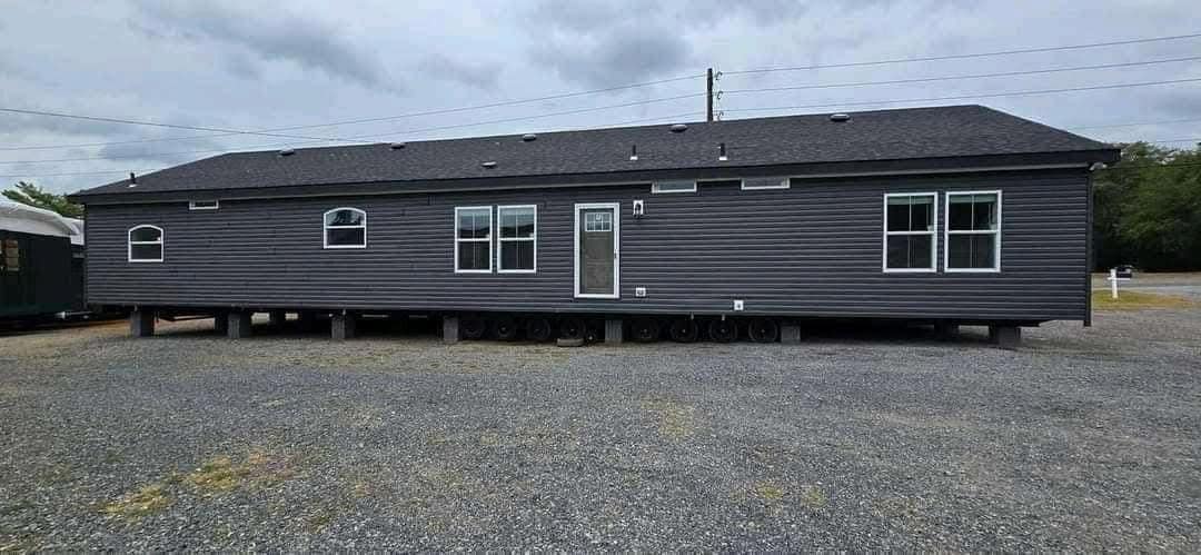 A gray mobile home with white-trimmed windows and several wheels underneath sits on a gravel lot under a cloudy sky, conveying a static and serene mood.