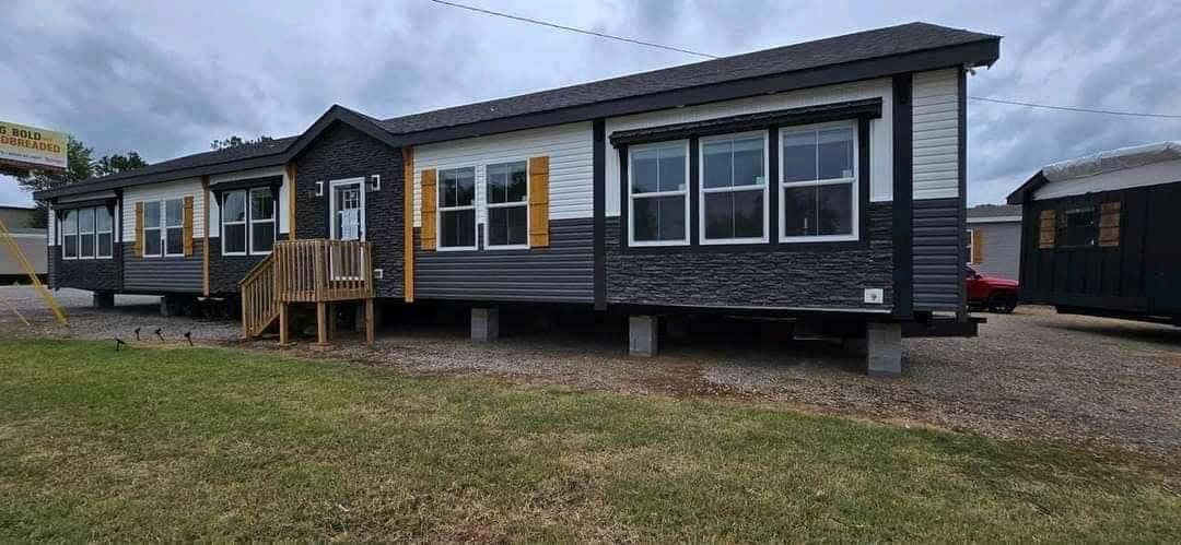 A modern, dark-toned manufactured home with white trim and yellow shutter accents sits elevated on a gravel lot. Overcast sky and grassy foreground.