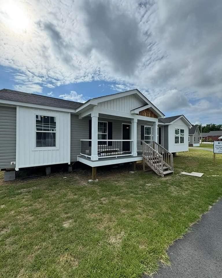 A modern, single-story prefabricated home with a porch and steps stands under a cloudy sky. The exterior features white and gray siding, creating a welcoming feel.