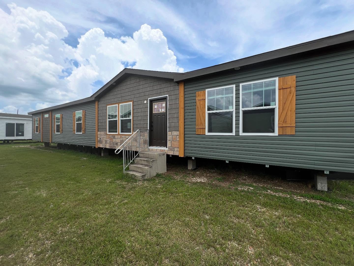 Gray modular home with wooden shutters and stone accents, set on a grassy lot under a bright, cloudy sky. Steps lead to a dark front door.