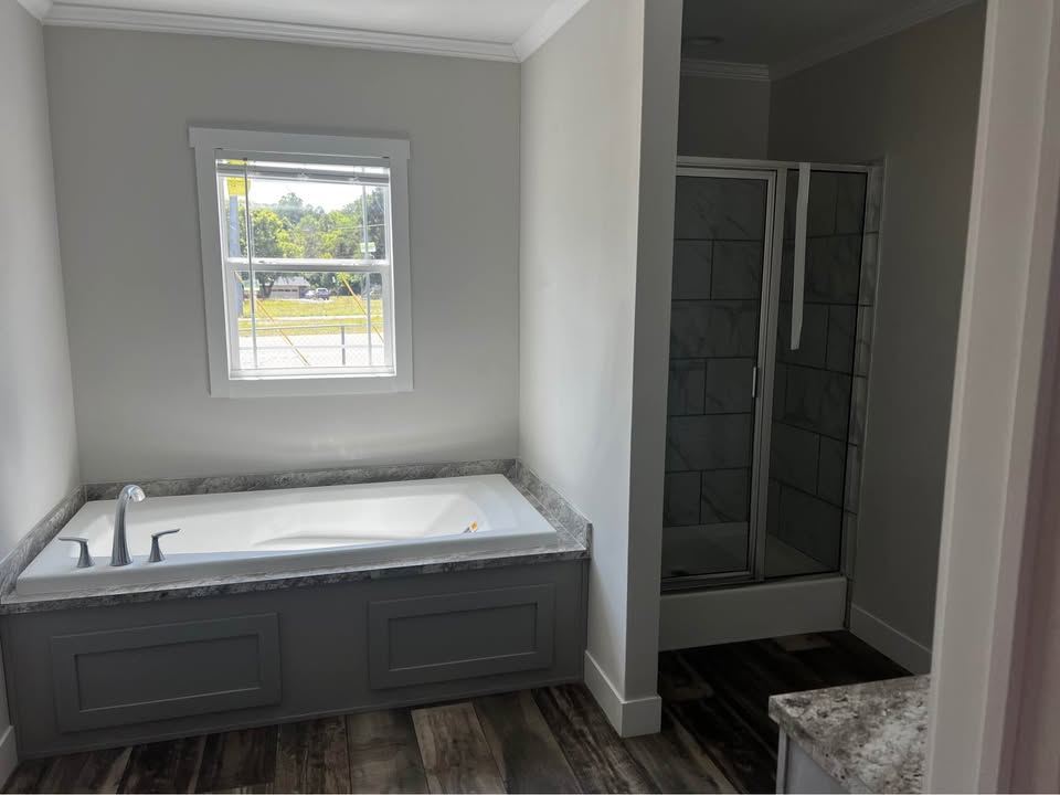 A modern bathroom with a large bathtub under a window, featuring a marble countertop. A glass-enclosed shower is on the right, with wood-patterned flooring.