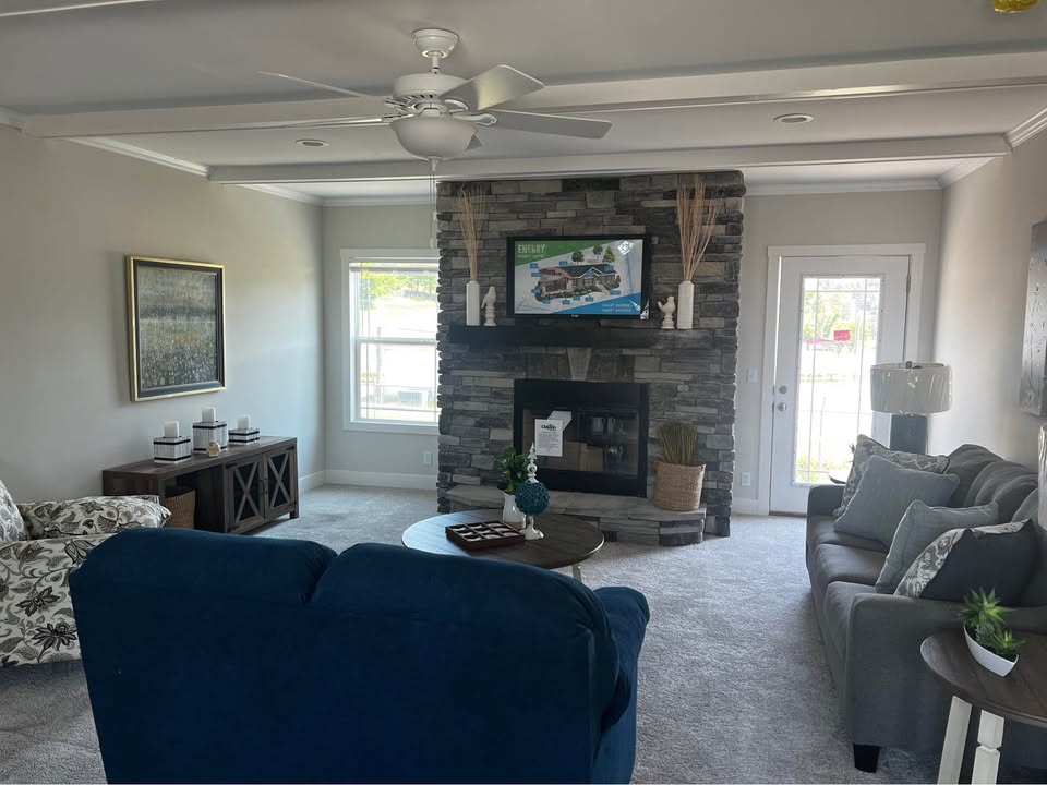 Cozy living room with gray stone fireplace, dark wood furniture, and blue and gray seating. Natural light pours in through large windows. Serene ambiance.