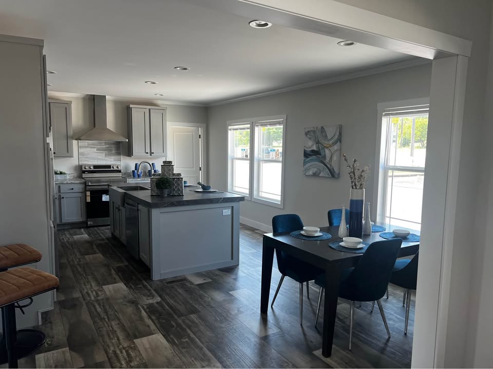 Modern kitchen and dining area with sleek gray cabinetry, an island, and elegant decor. Natural light streams through large windows, creating an inviting ambiance.