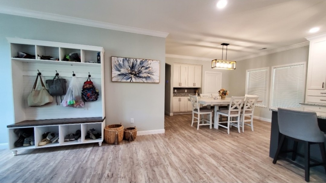 A modern, light-filled dining area with a wooden floor. A wall with hooks and shelves for storage is on the left. A white table, chairs, and two woven baskets add a cozy touch.