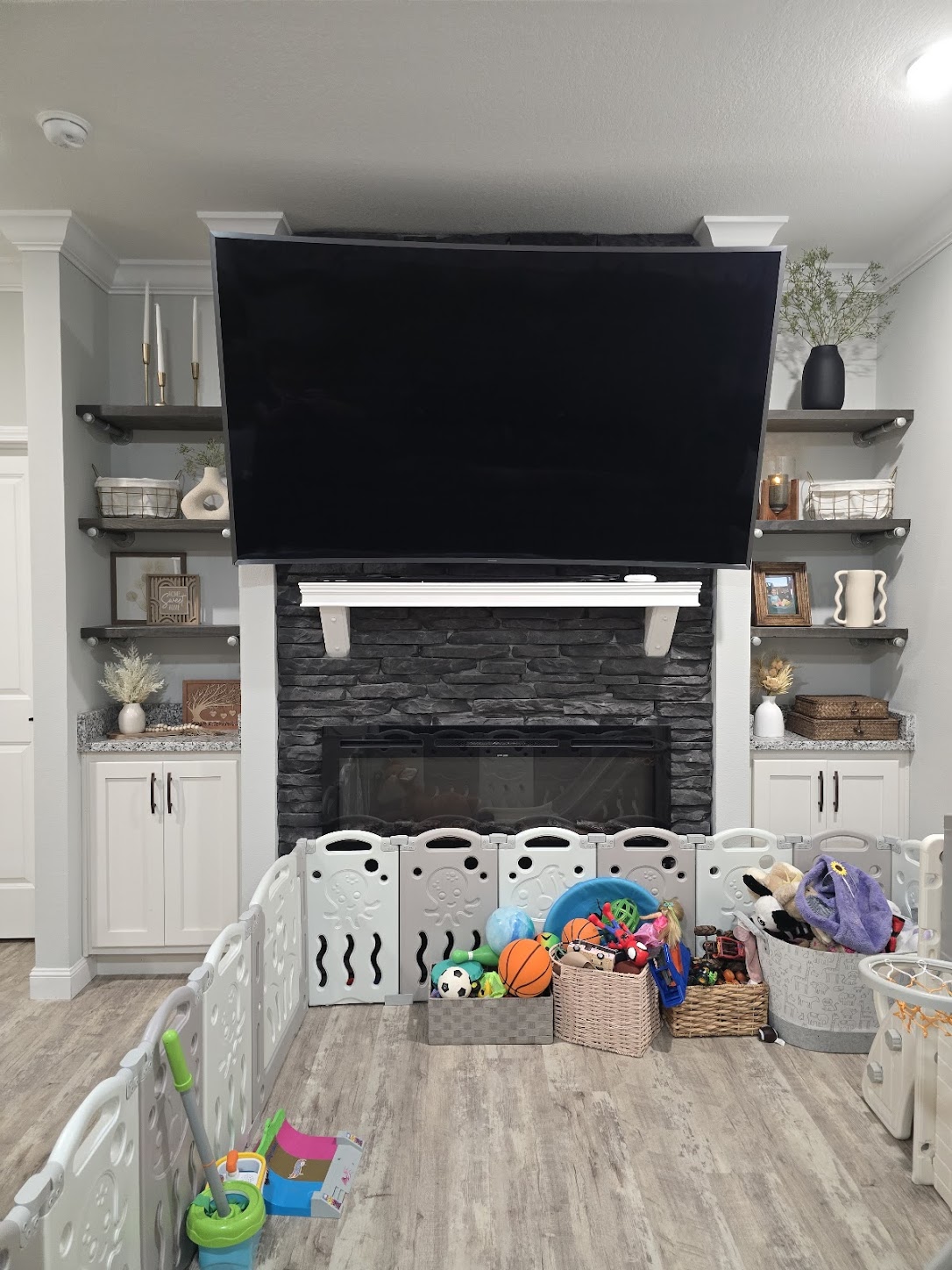 Family room with a wide-screen TV above a stone fireplace, flanked by shelves with decor. A play area has toy storage and a safety gate. Cozy and organized.
