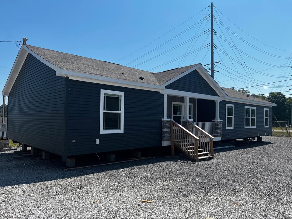 A dark blue modular home sits on a gravel lot under a clear blue sky. It features a small front porch with stairs and several windows.