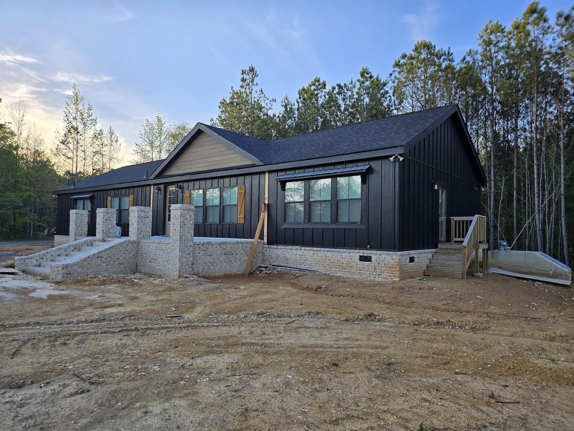 Modern black barn-style house with large windows, wooden shutters, and brick foundation set against a backdrop of trees and clear blue sky.