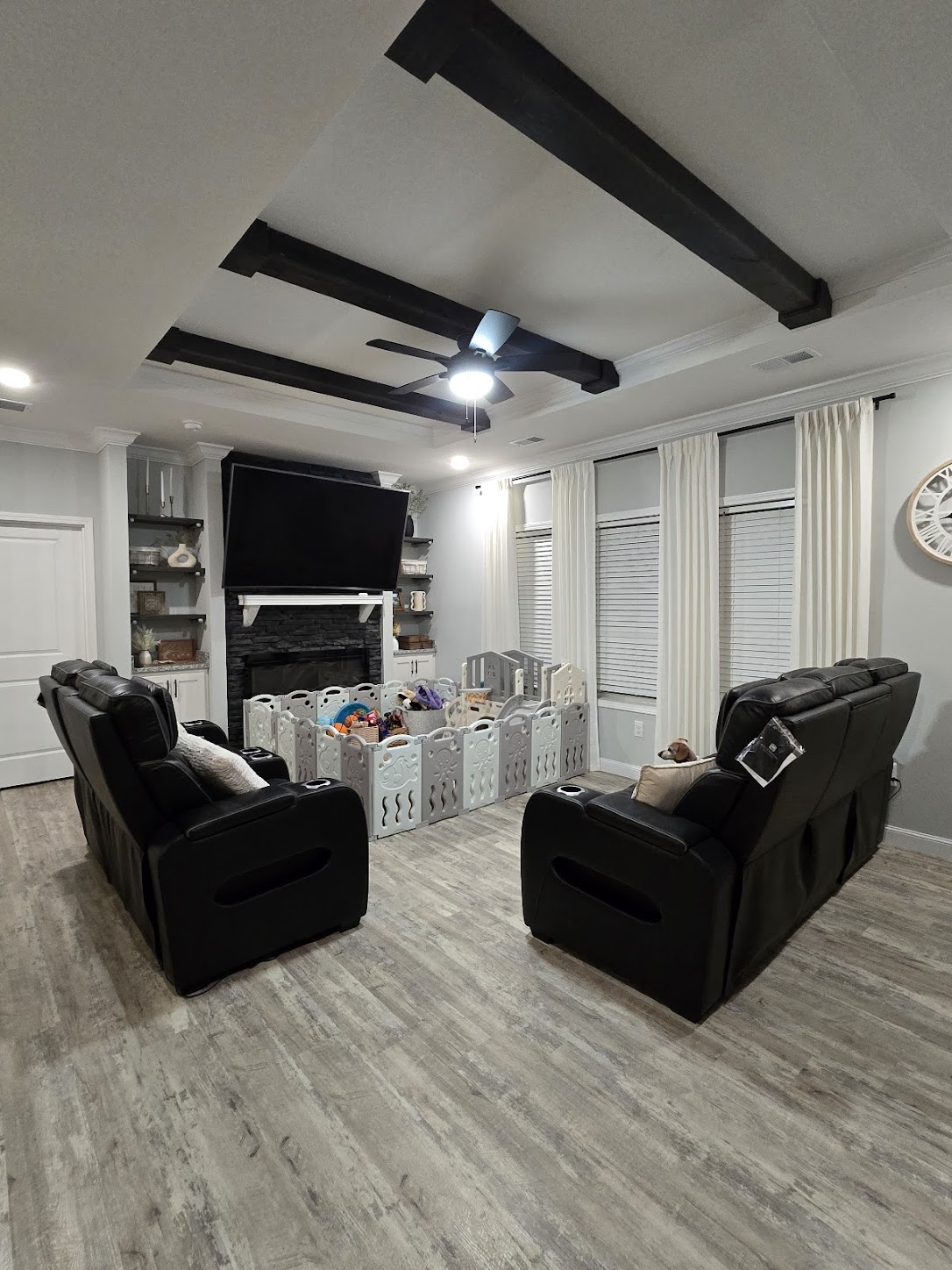 Spacious living room with grey flooring, black recliners, and a baby playpen filled with toys. Mounted TV above a dark fireplace, bright curtains, and modern decor.