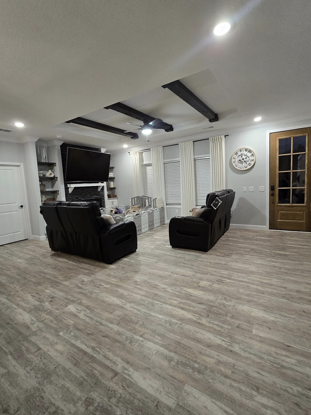 A cozy living room with wood flooring, two black recliners facing a wall-mounted TV above a fireplace, white drapes, and a rustic wall clock.