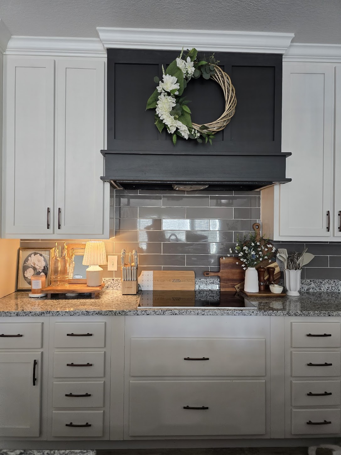 A modern kitchen with white cabinets and a gray backsplash. A floral wreath hangs above the stove. Counter features a lamp, knife set, and decor.