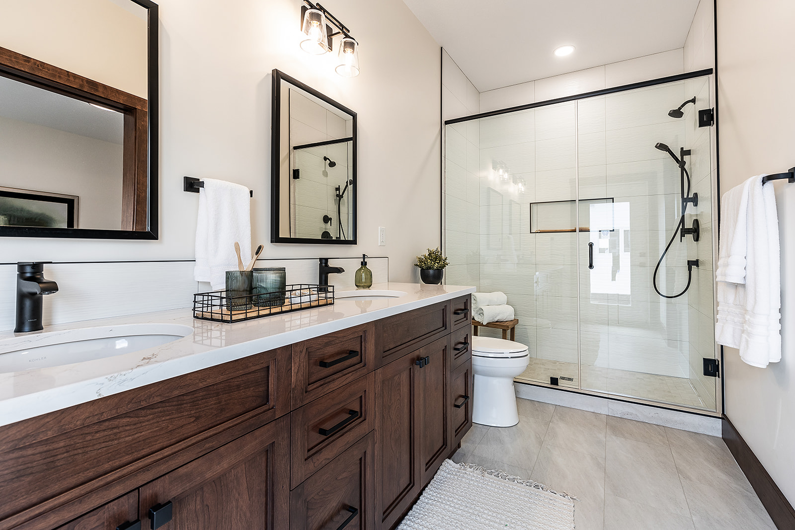 Elegant bathroom with a wooden vanity, marble countertop, and dual sinks. Glass shower enclosure with black fixtures. Soft, warm lighting and neutral tones.