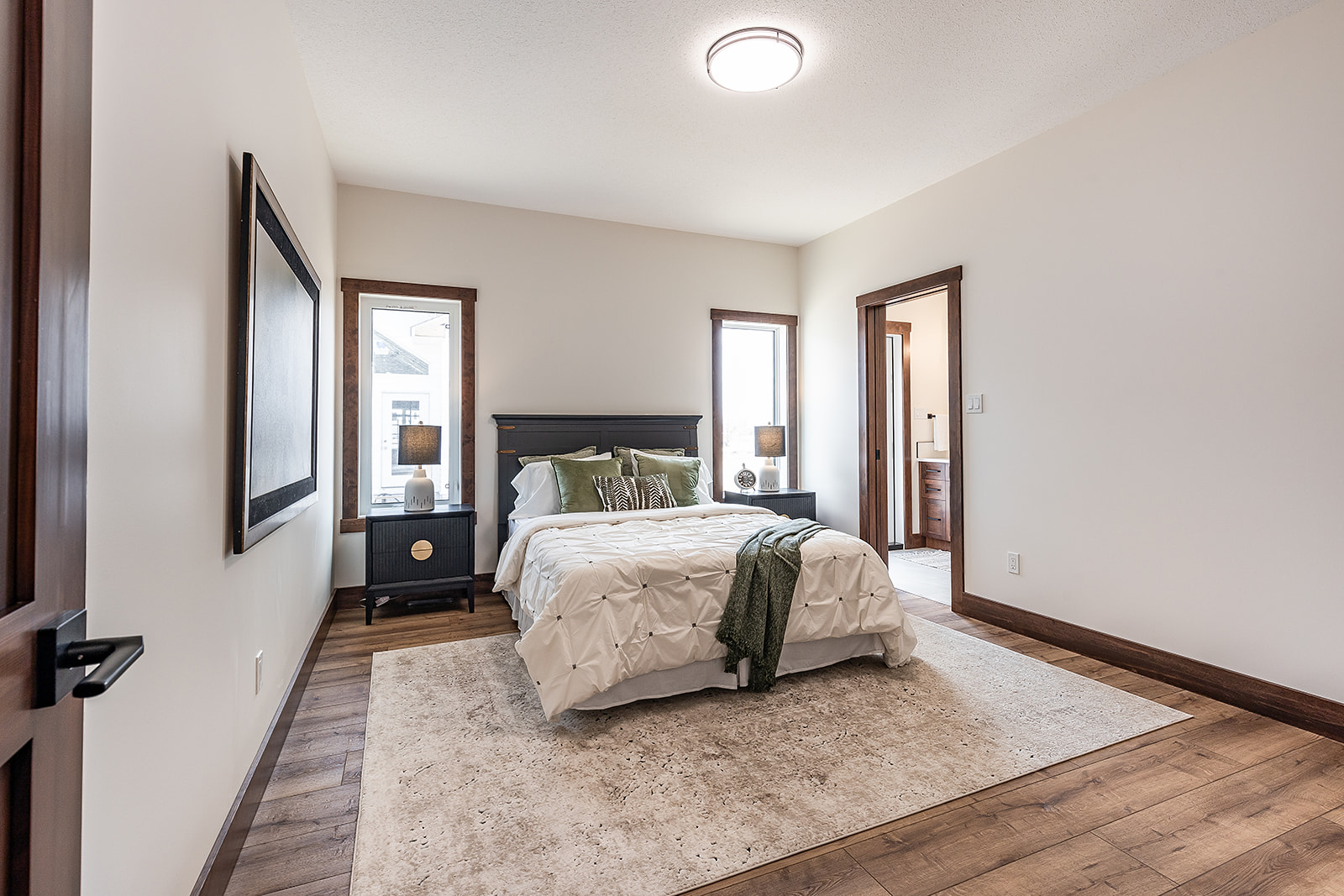 A modern bedroom with a neatly made bed featuring green and white pillows and a textured blanket. Wooden furniture and floor create a warm, inviting tone.