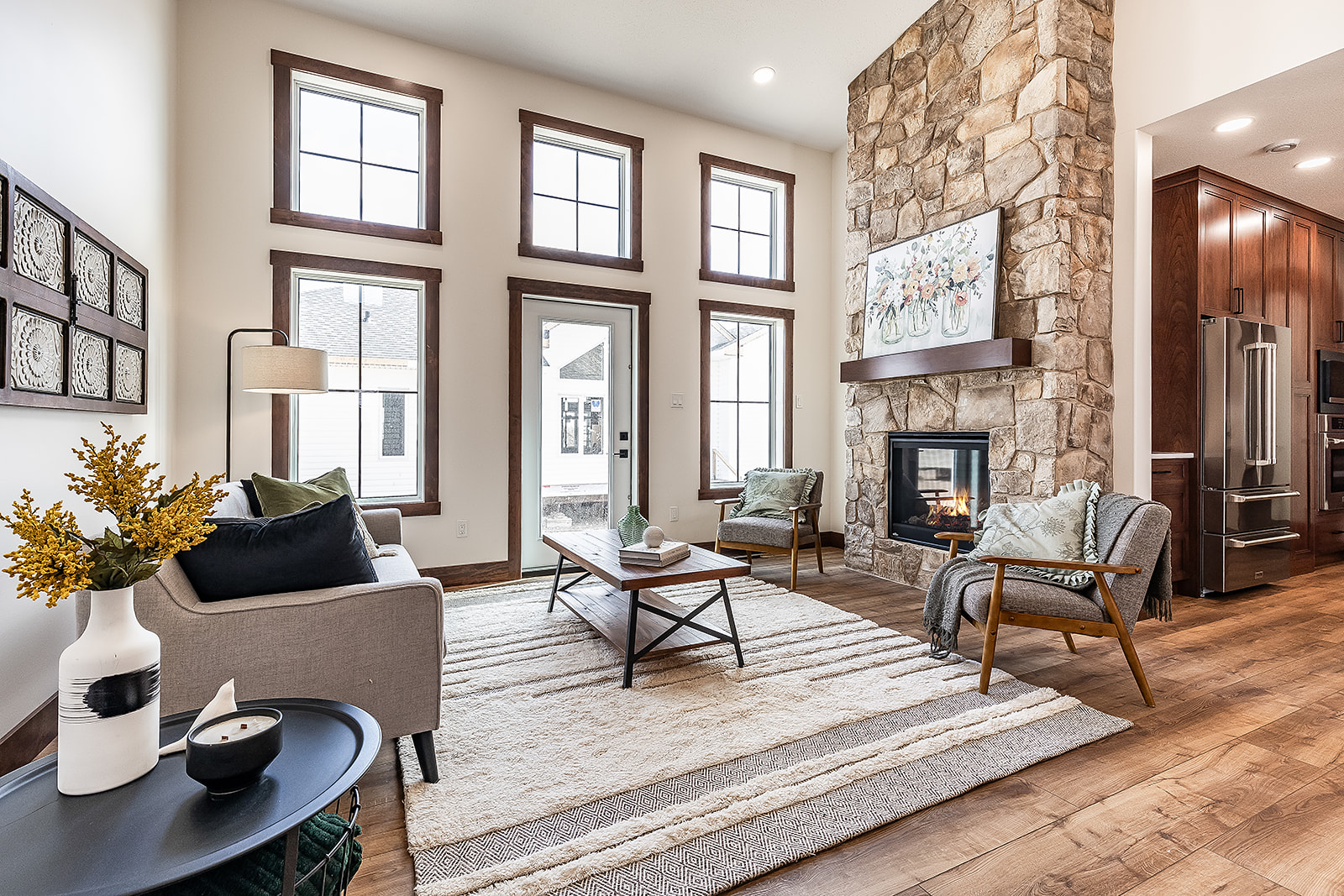 Bright, modern living room with tall windows, stone fireplace, and cozy seating. Wooden floors and earth tones create a warm, inviting atmosphere.