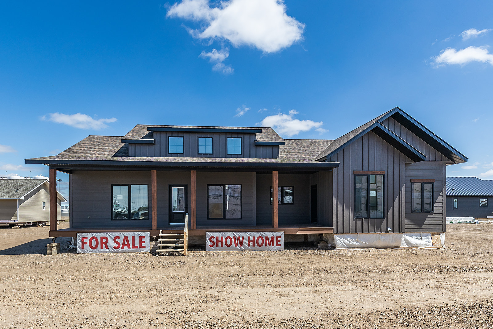Single-story gray show home with gable roof, wooden porch, and "For Sale" sign. Bright blue sky with clouds adds a welcoming tone.