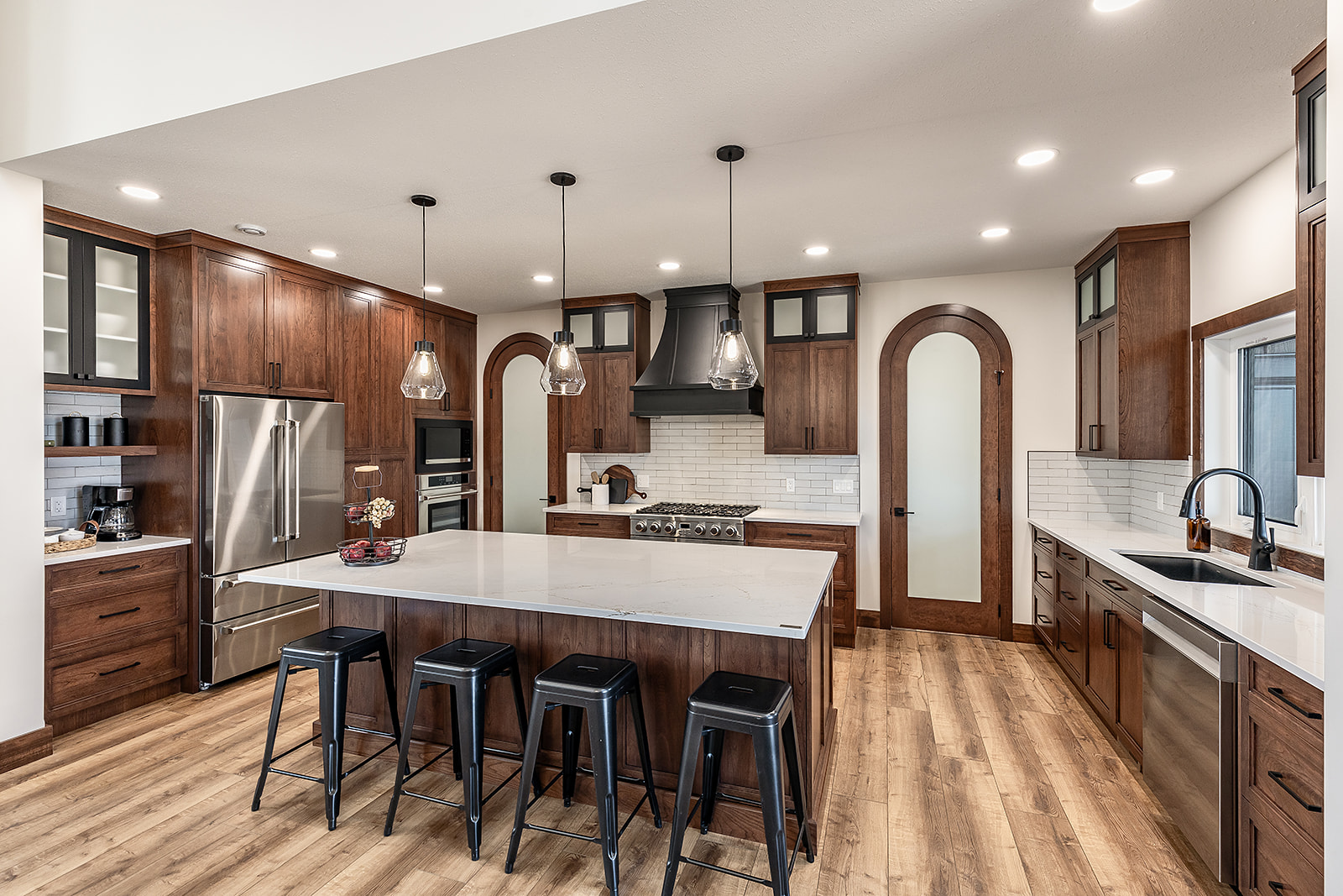 Modern kitchen with wooden cabinets, marble island, and black stools. Pendant lights hang above the island, creating a warm, sophisticated feel.