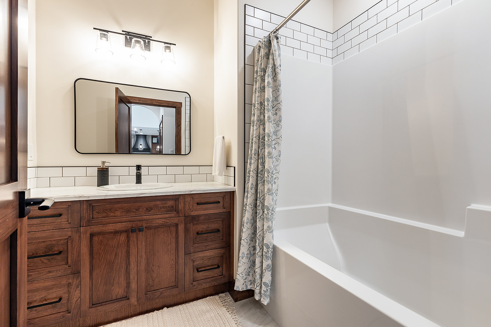 Modern bathroom with wood cabinet vanity, white countertop, rectangular mirror, and wall lights. White tub with patterned curtain; calm ambiance.
