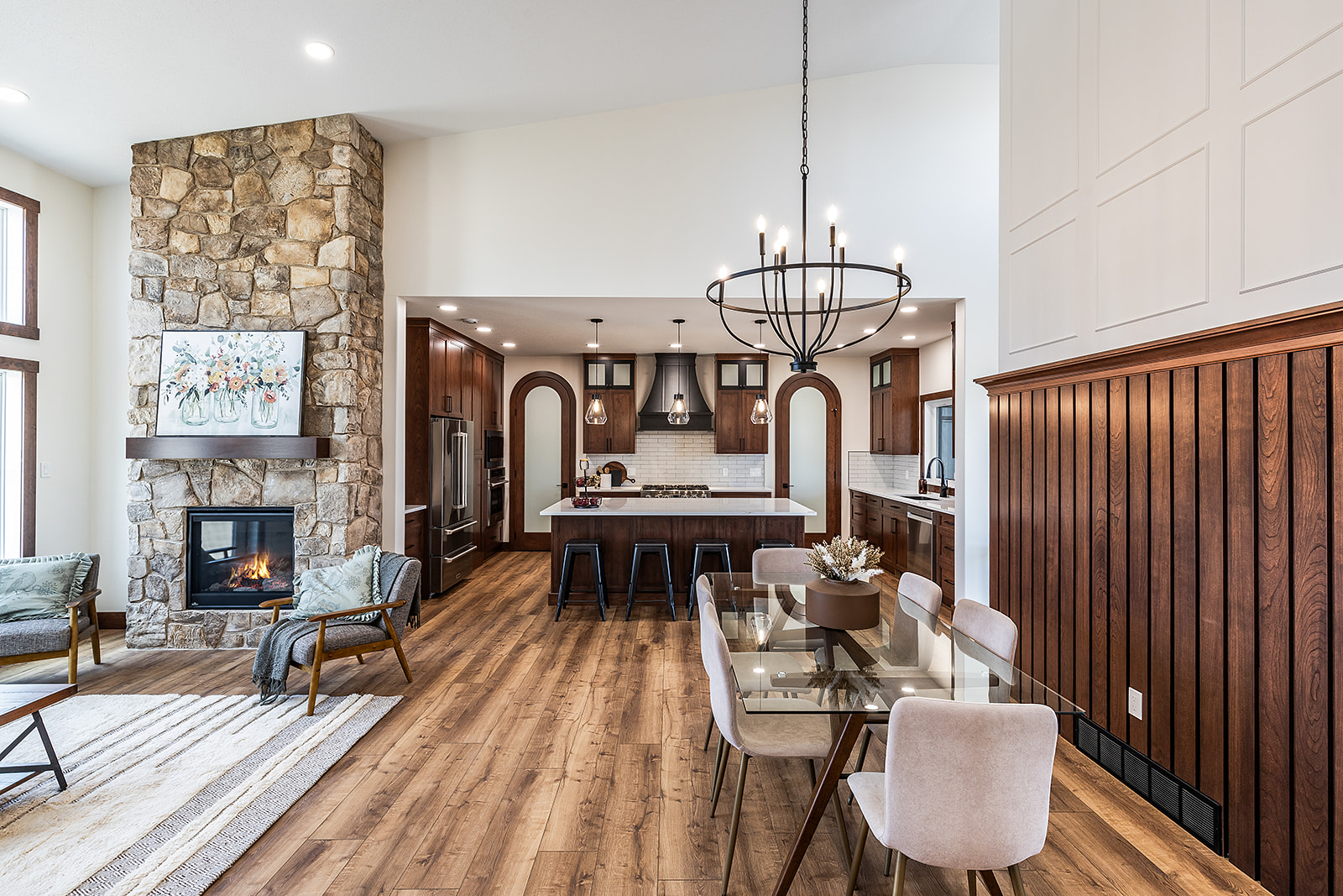 Spacious, elegant open-plan living area with a stone fireplace, cozy chairs, a modern kitchen, and a glass dining table. Warm wood tones and soft lighting create an inviting atmosphere.