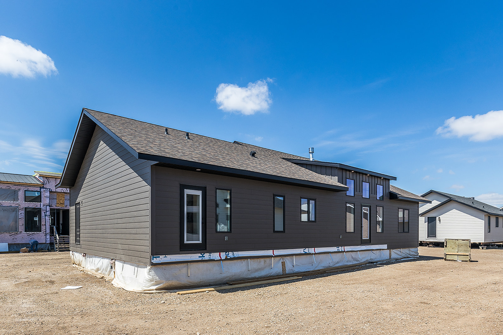 A newly constructed modular home on a clear day. The house features dark siding, multiple windows, and a shingled roof, with a bright blue sky above.