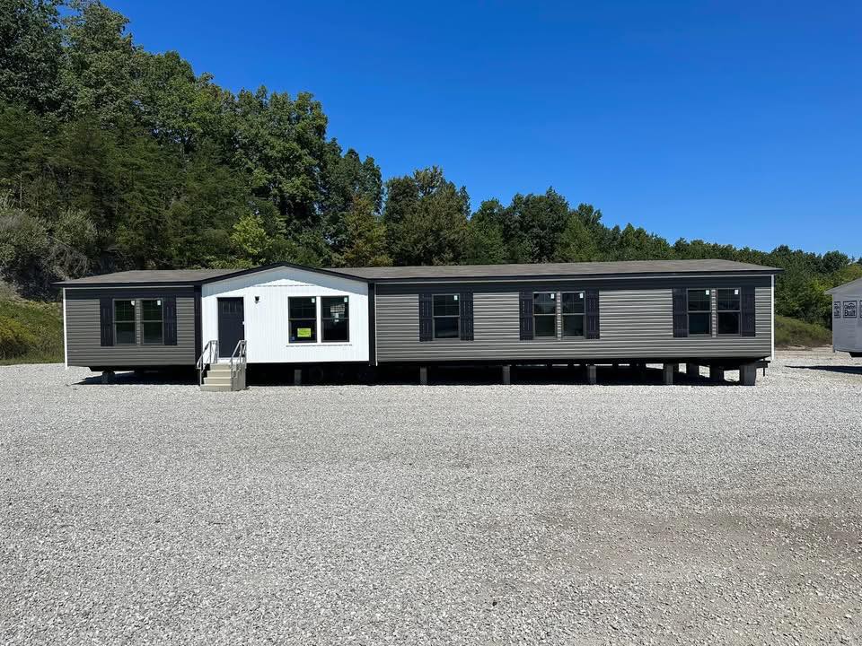 A modern, double-wide manufactured home with gray siding and black shutters sits on a gravel lot surrounded by trees under a clear blue sky.