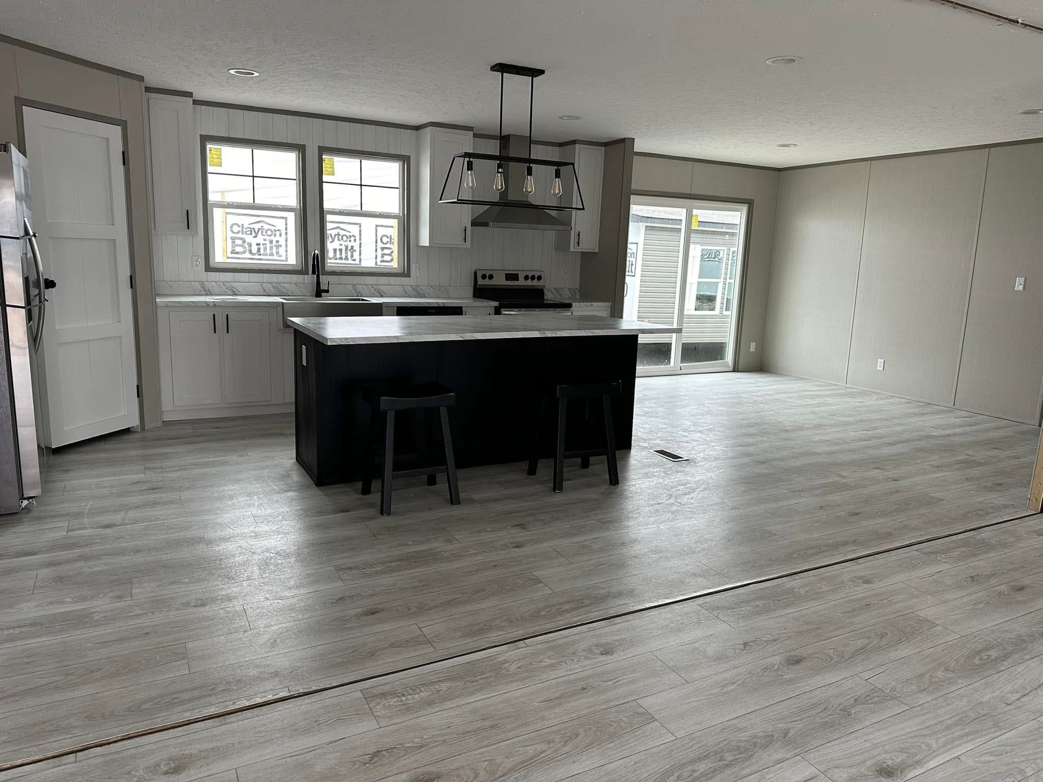 Spacious kitchen with light wood flooring, featuring a central island with two stools, modern pendant lighting, and sleek white cabinetry under large windows.