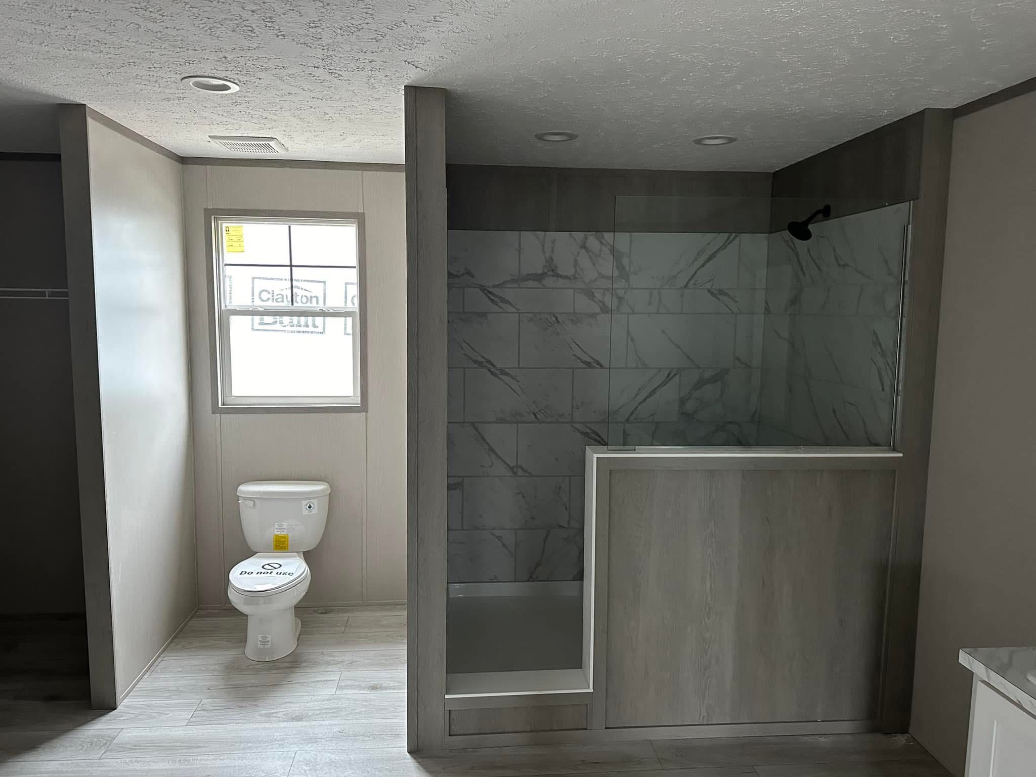 A bathroom with a modern shower on the right featuring marble tiles and a glass partition. A toilet is on the left under a window, creating a minimalistic and clean atmosphere.