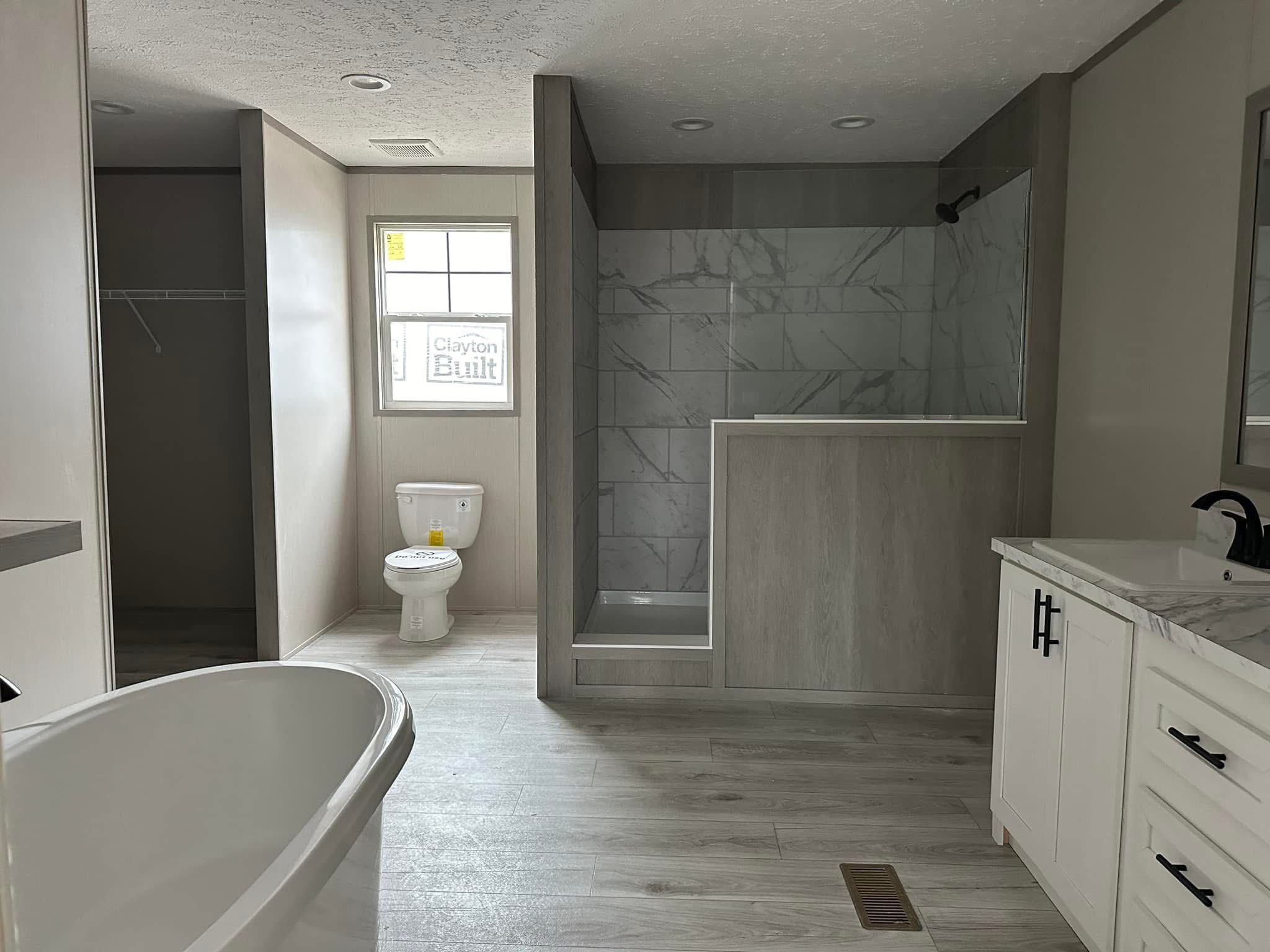 Modern bathroom with a freestanding bathtub, marble-tiled shower, white vanity, and toilet near a window. The light wood floor adds an airy feel.