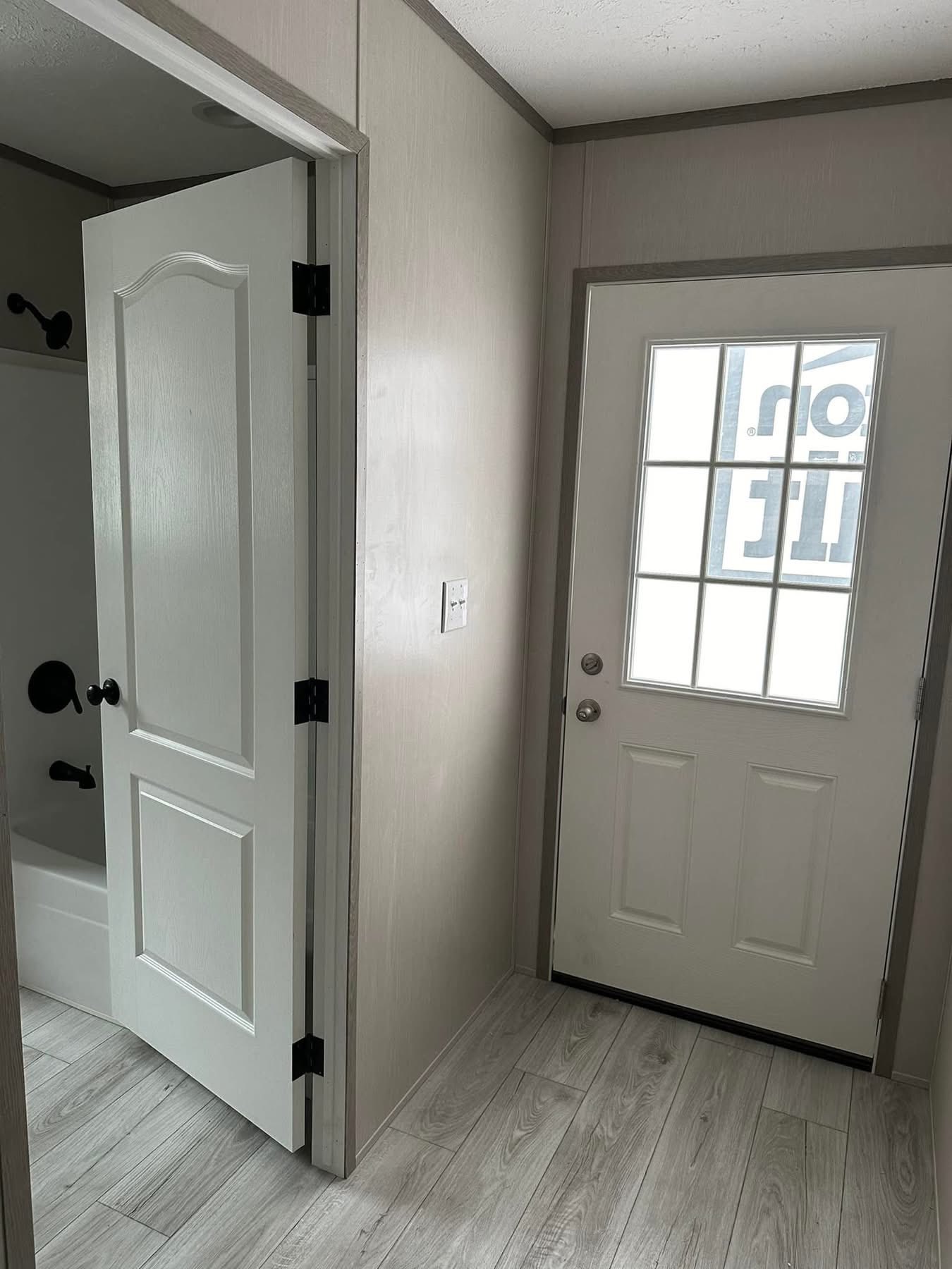 Interior entryway with light wood flooring. A white door with glass panels is at the right, and a partially open door to a bathroom on the left. The space feels clean and minimal.