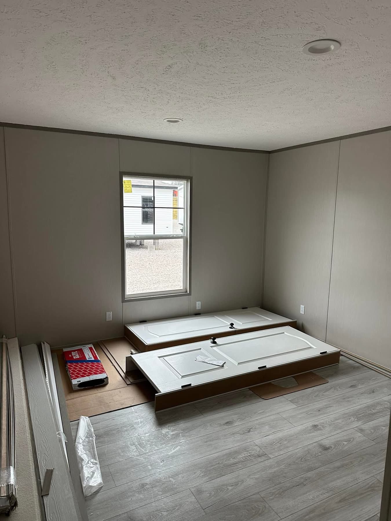 Empty room under construction with gray walls and flooring. A window shows a view outside. White doors and construction materials are on the floor. Calm atmosphere.