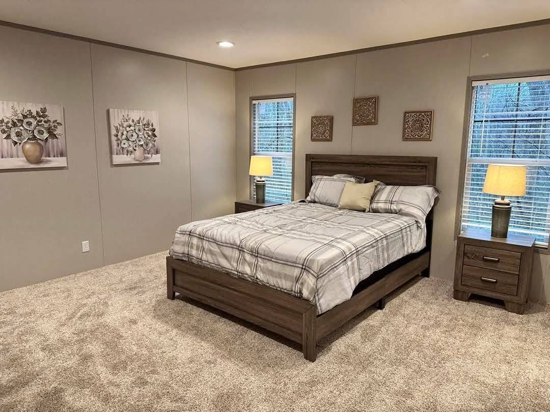 A cozy bedroom with a neatly made bed featuring plaid bedding and a beige accent pillow, flanked by two windows with blinds and wooden nightstands, each topped with a lit lamp. Light gray carpet and neutral walls adorned with floral paintings and decorative panels contribute to a calm, inviting atmosphere.