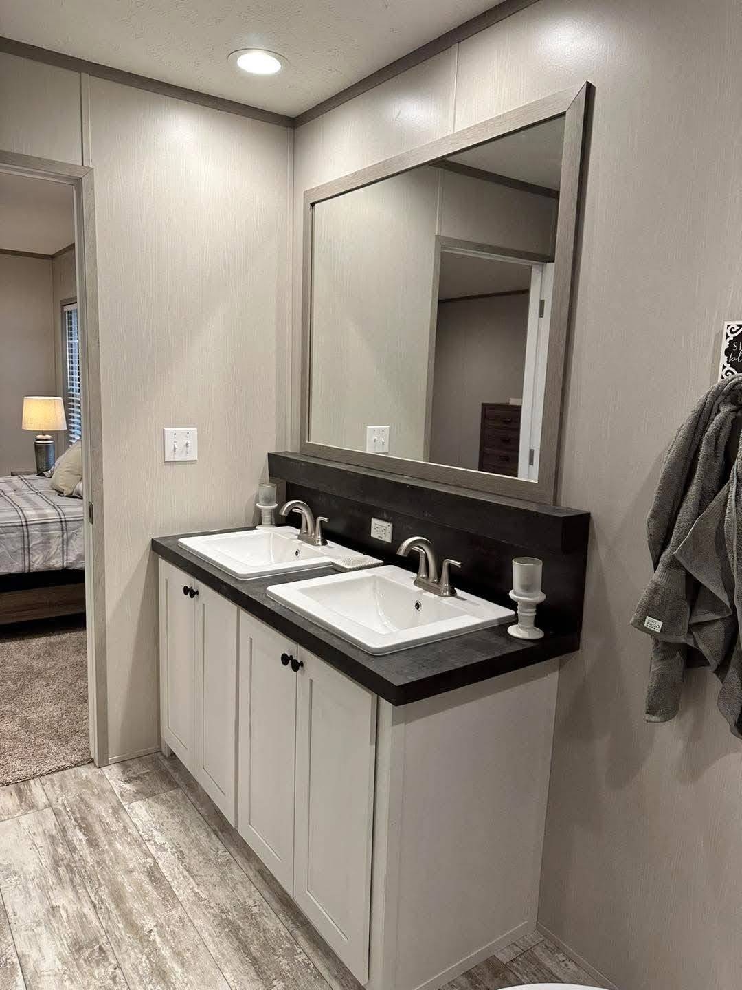 Modern bathroom with double sinks on a dark counter, large mirror, and minimalist design. Soft lighting adds warmth. Bedroom visible through open door.