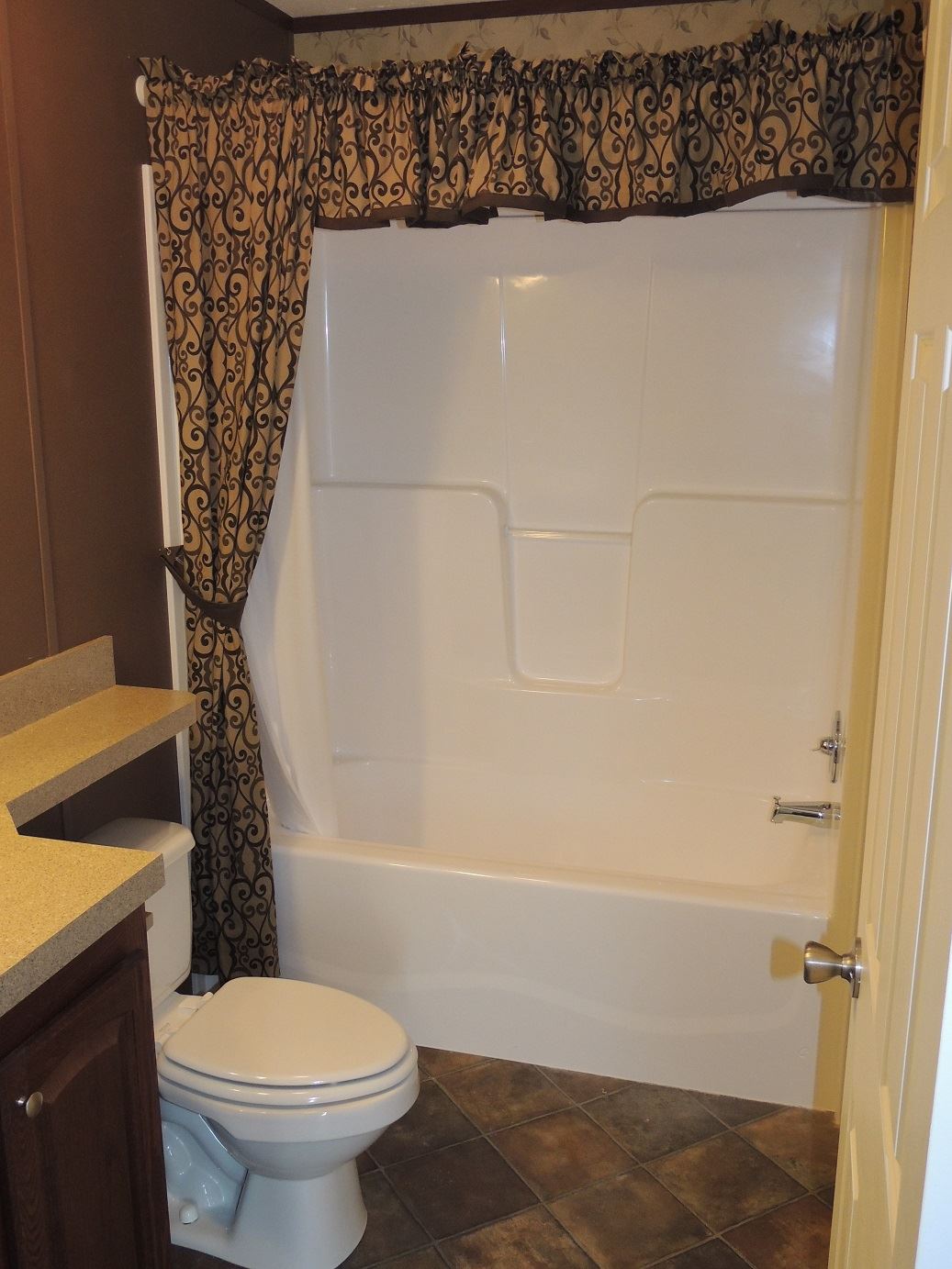 A compact bathroom featuring a white bathtub with ornate brown curtains, a white toilet, beige countertop, brown cabinets, and tiled floor.