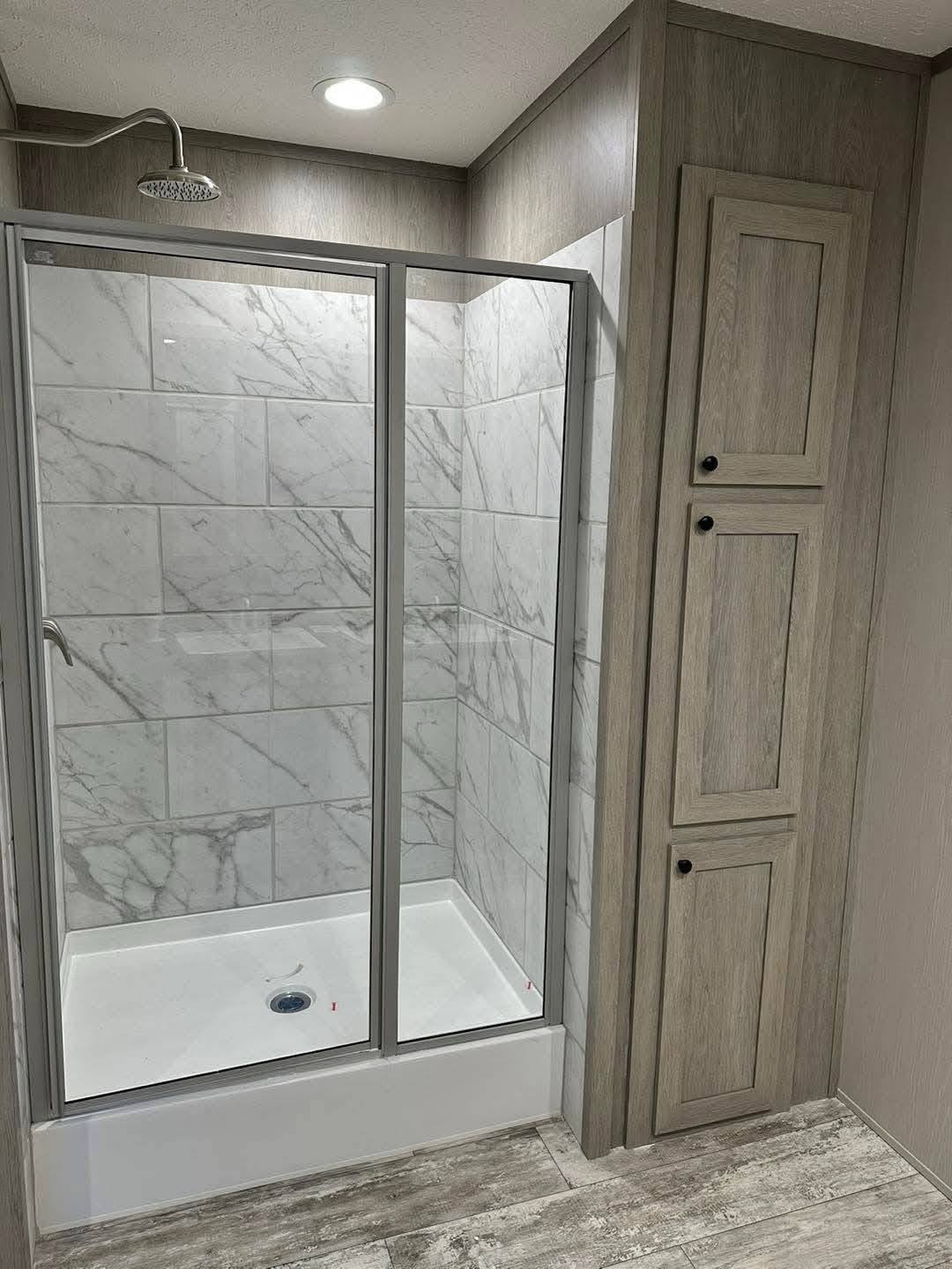 Modern bathroom with a glass-enclosed shower. The walls feature gray marble tiles. There's a wooden cabinet with two doors beside the shower. Cozy, minimalist vibe.