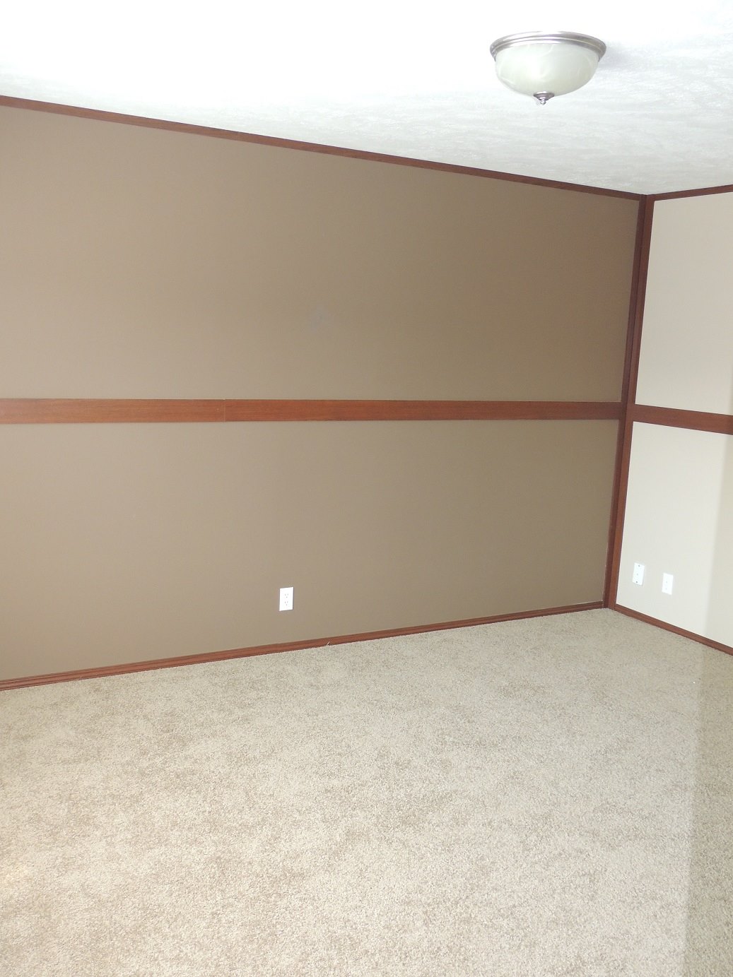 An empty room with beige walls and carpet, featuring wood trim and a ceiling light. The room has a clean, minimalist atmosphere.
