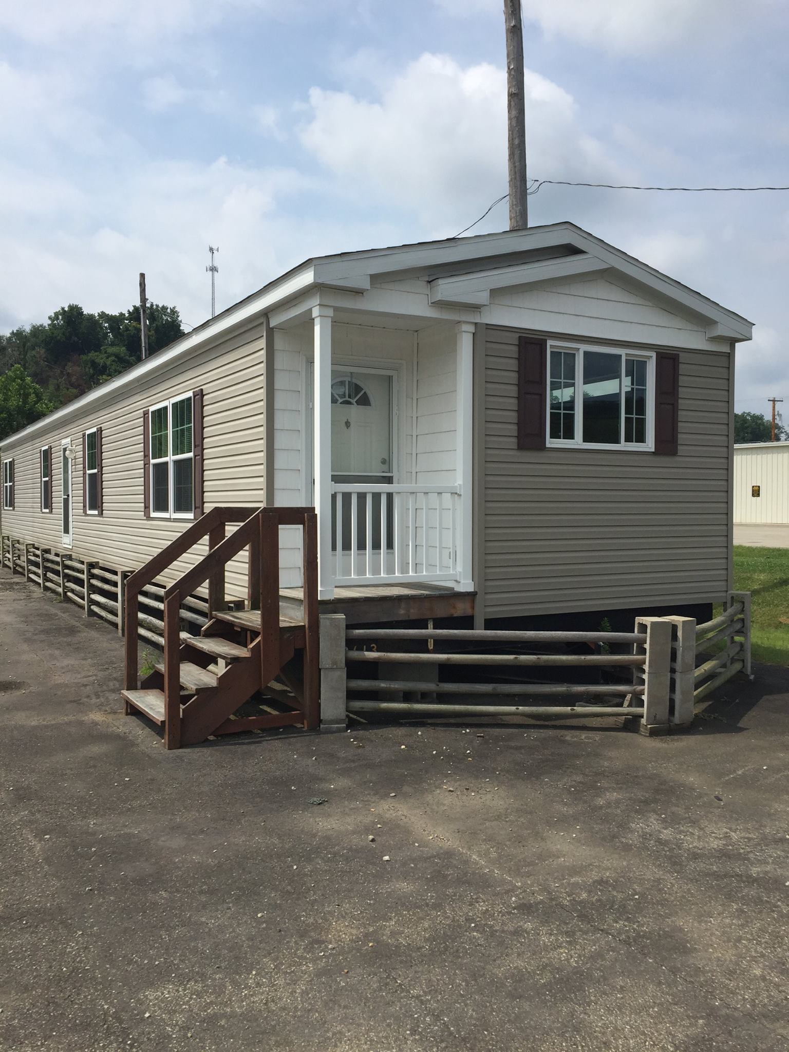 A beige and white mobile home with a small porch and wooden stairs is shown under a partly cloudy sky. A wooden fence lines the front.