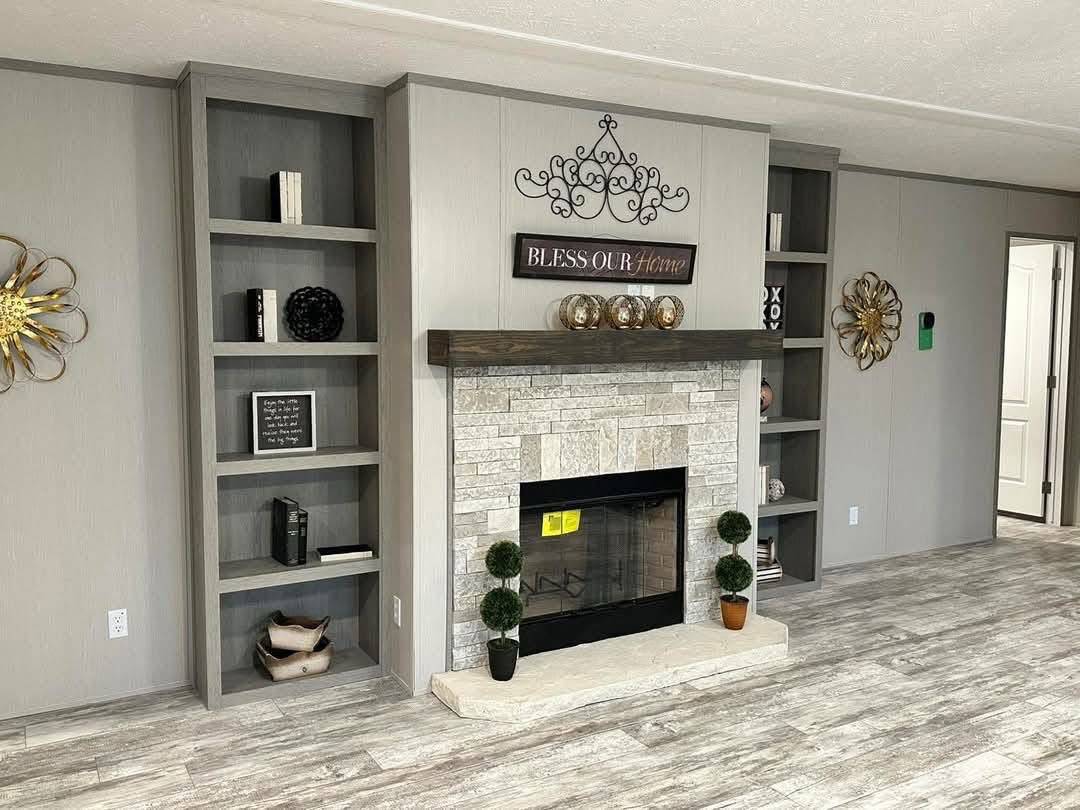 Modern living room with a stone fireplace, flanked by gray bookshelves. Decor includes metal wall art, a “Bless Our Home” sign, and potted plants.