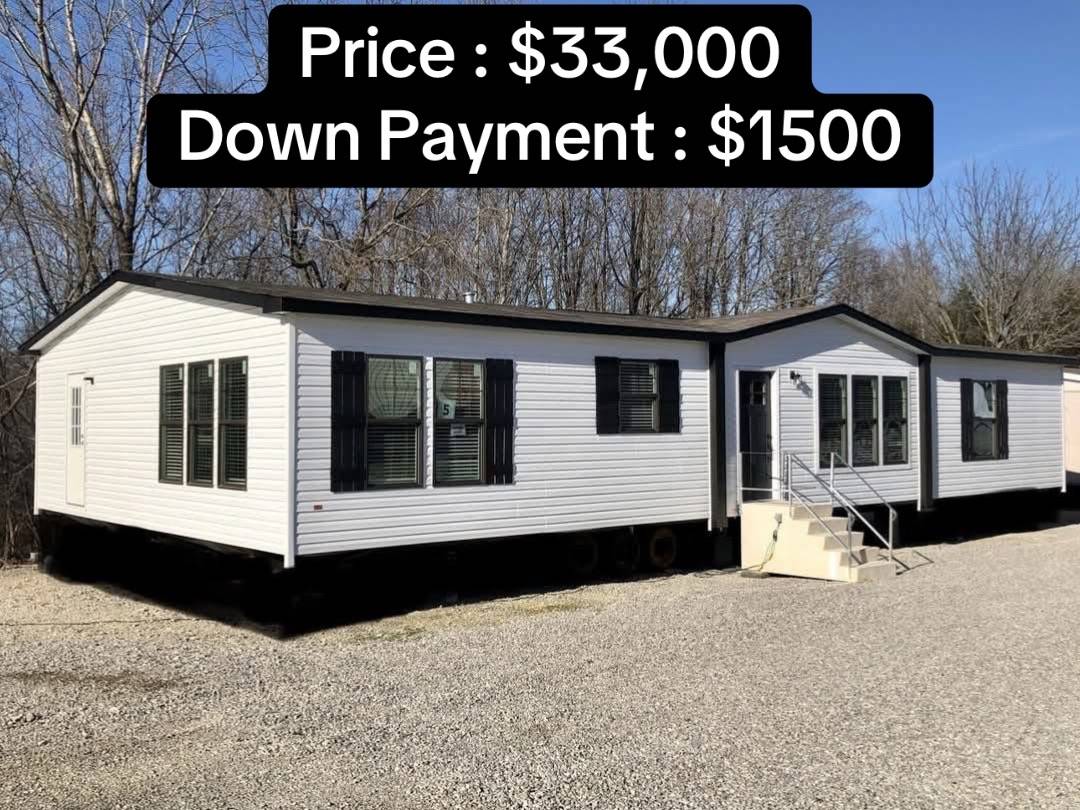 White mobile home on gravel lot with trees in the background. Text overlay shows price: $33,000 and down payment: $1,500. Bright and clear day.