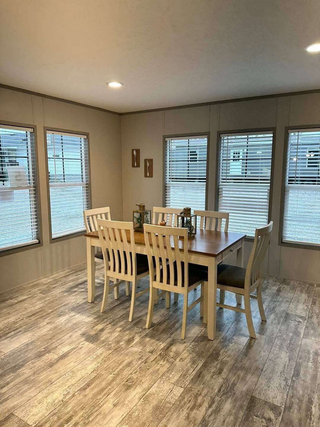 A cozy dining room with a wooden table set for six under soft lighting. Three windows with blinds diffuse natural light onto wood-patterned floors.