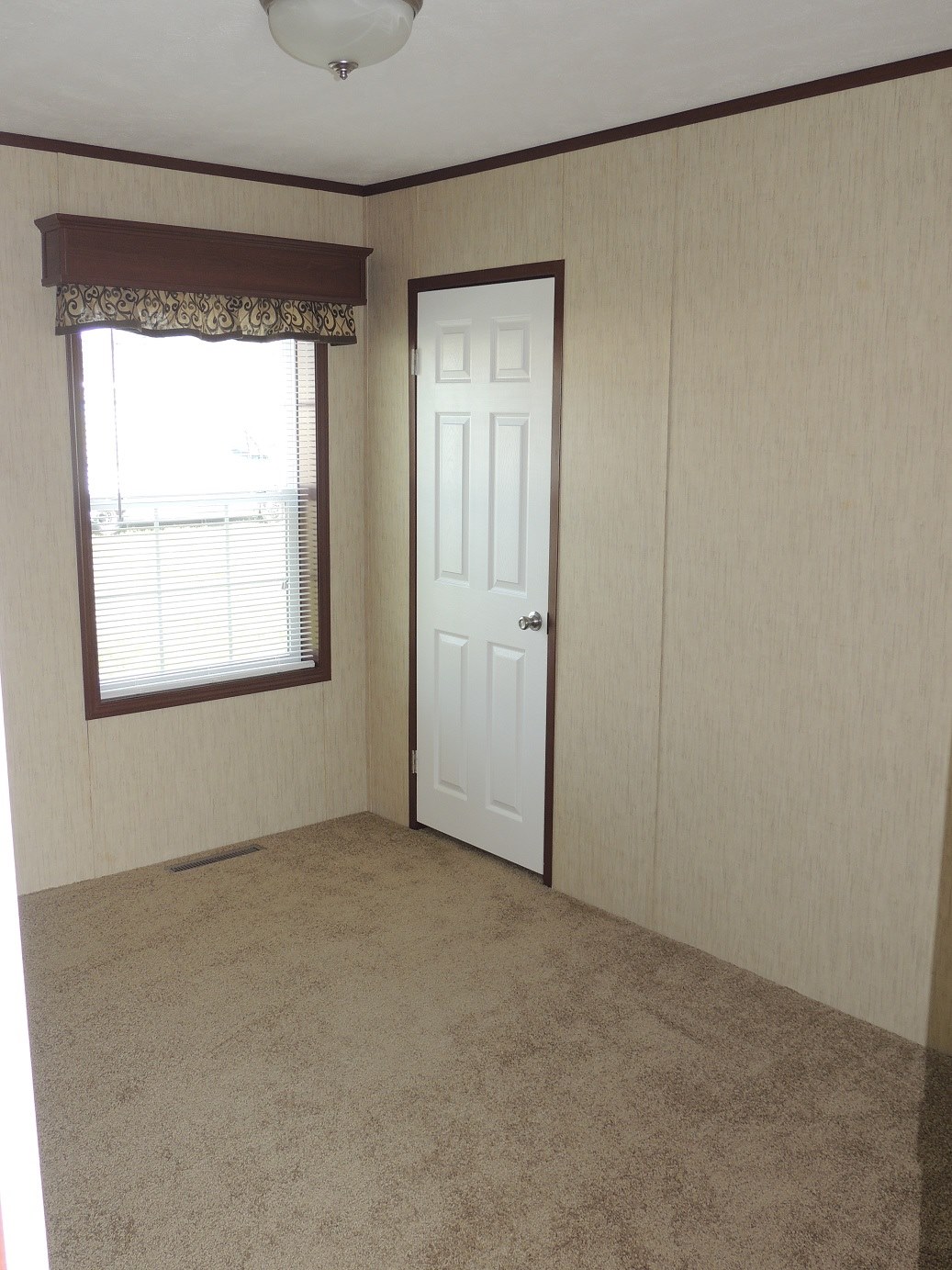 A small, empty room with beige carpet and light beige walls. A window on the left has closed blinds and a brown valance. A closed white door is on the right.