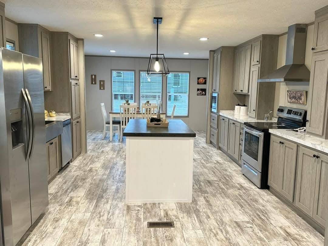 Modern kitchen with wooden cabinets, stainless steel appliances, a central island with dark countertop, and a cozy dining area in the background.