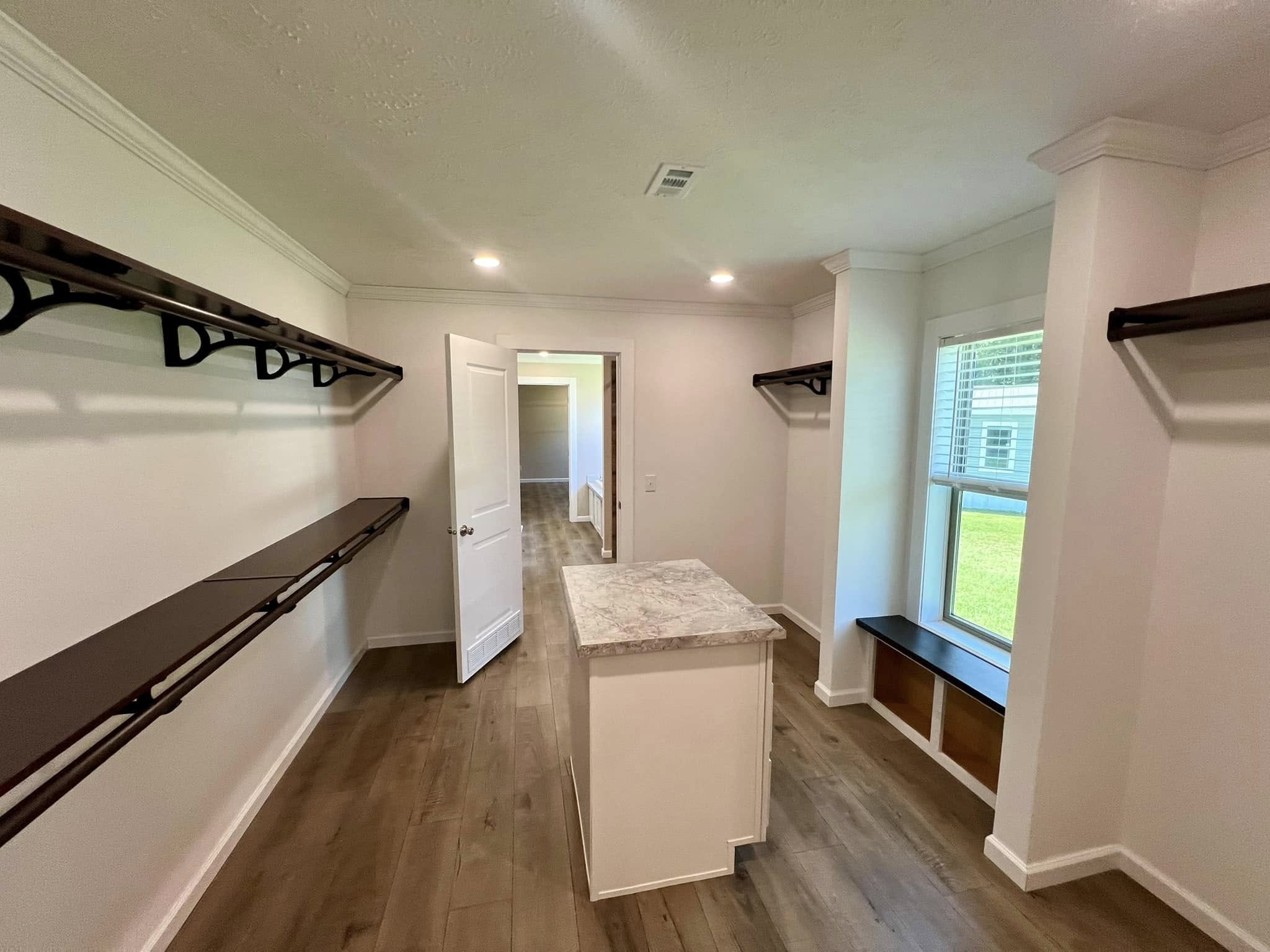 A spacious, well-lit walk-in closet with wooden shelves on white walls, marble-topped island, and large window seat, evoking a sense of modern elegance.
