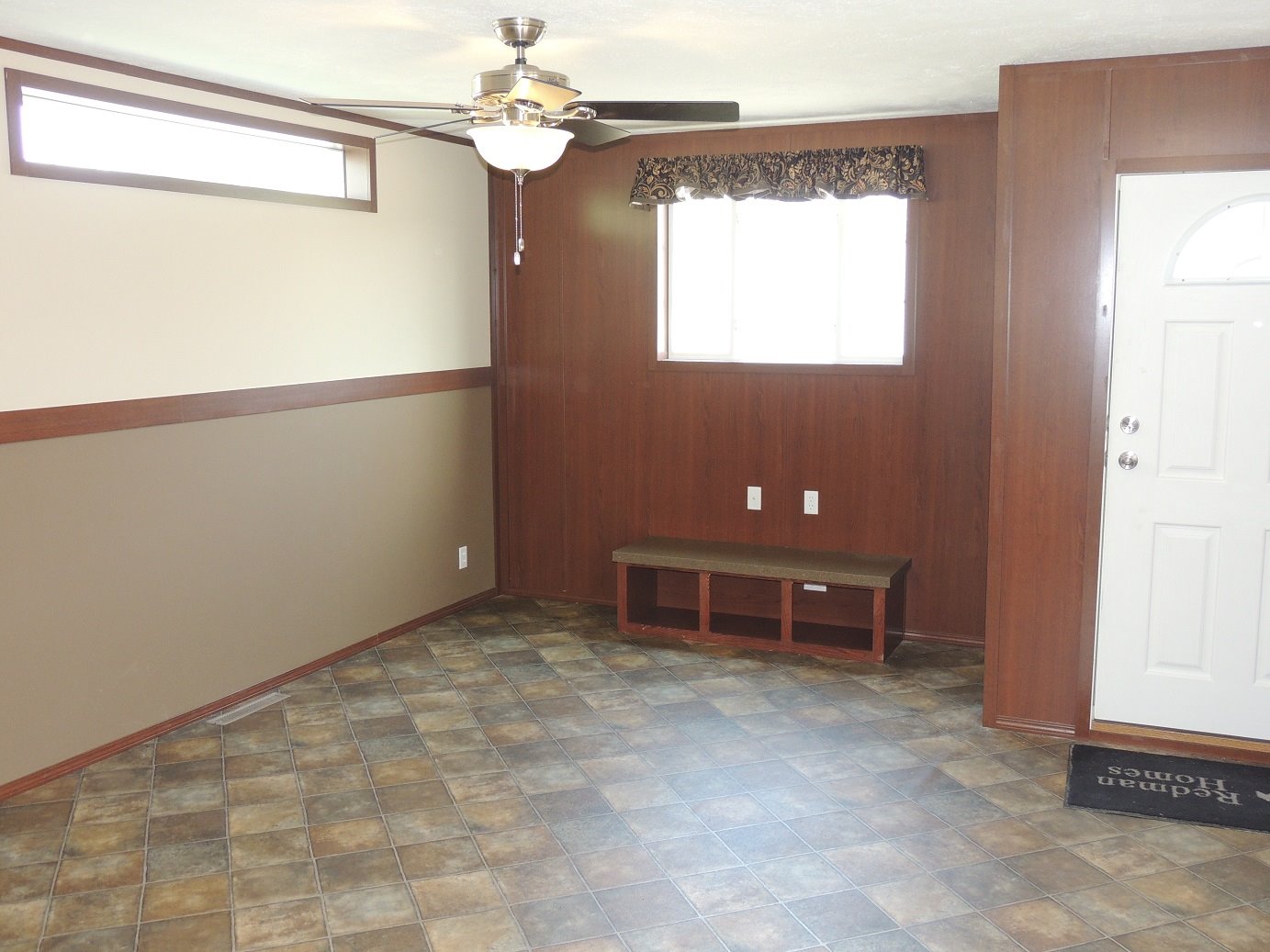A small, empty room with wood-paneled walls and a tiled floor. It features a ceiling fan, a small window with a curtain, and a white door.