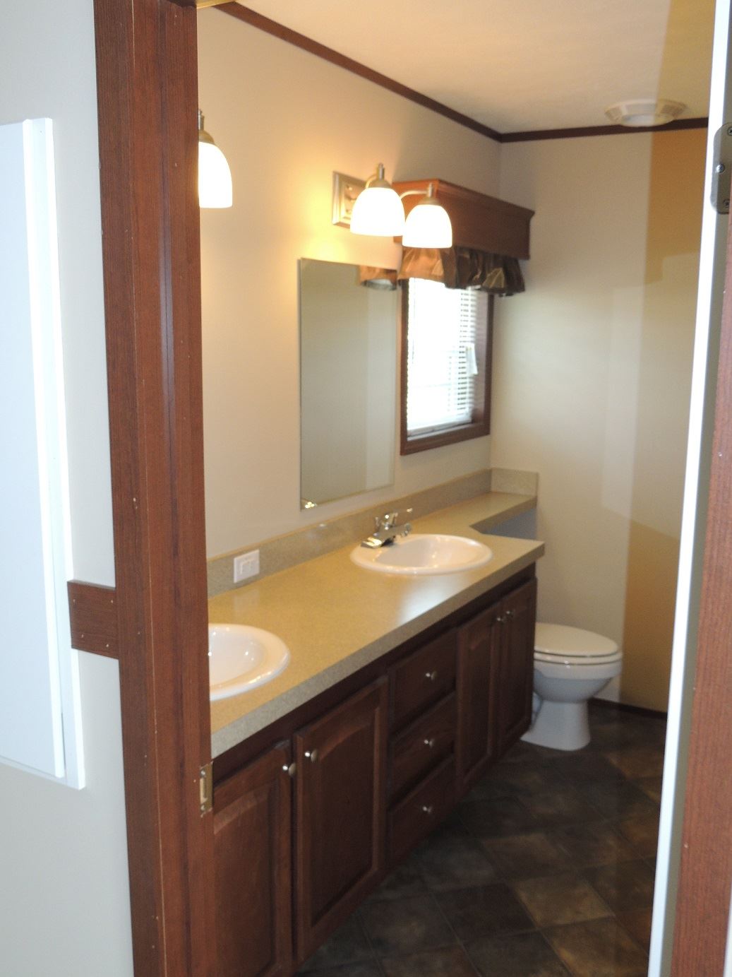 A cozy bathroom with a double sink vanity, large mirror, and soft lighting. Visible are a window with blinds and a toilet, creating a clean, functional space.