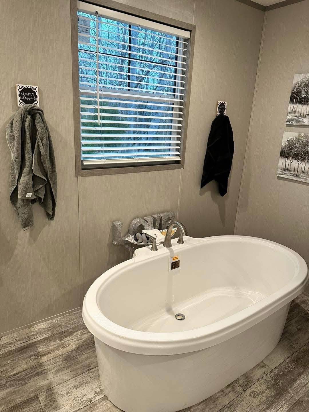 A modern bathroom with a white oval bathtub, gray-green walls, and a window with blinds. Towels hang nearby, creating a serene, cozy atmosphere.