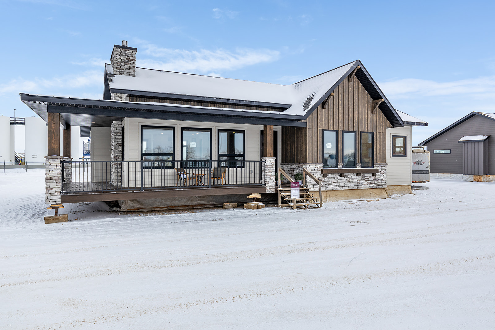 Modern cabin with snow-covered roof, large windows, and wooden panels. The porch features two chairs, with a calm, wintry backdrop and clear sky.