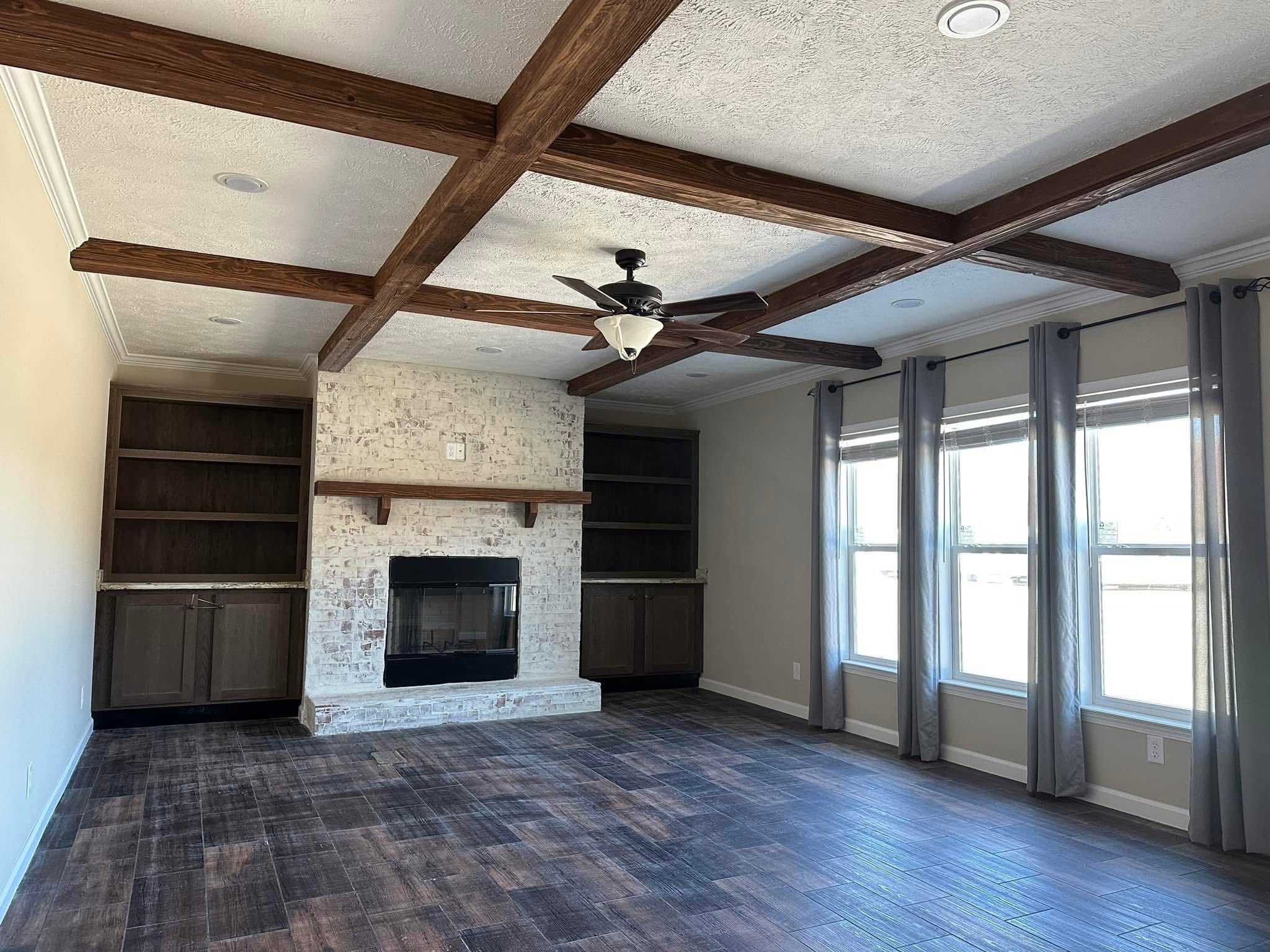Spacious living room with a rustic feel, featuring a wooden beam ceiling, brick fireplace, dark wood shelves, large windows, and hardwood floors.