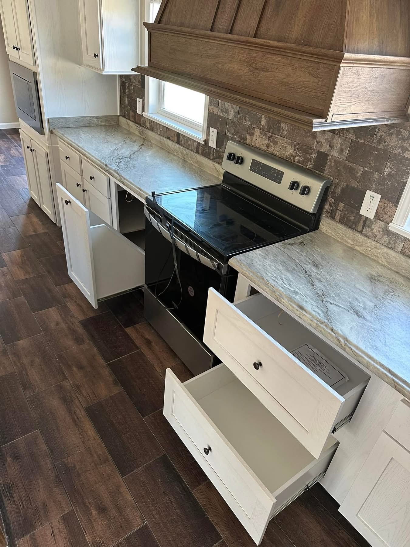 Modern kitchen with wood flooring, open white cabinets and drawers, black stove, gray marbled countertops, and a wooden range hood, conveying a sleek and functional design.